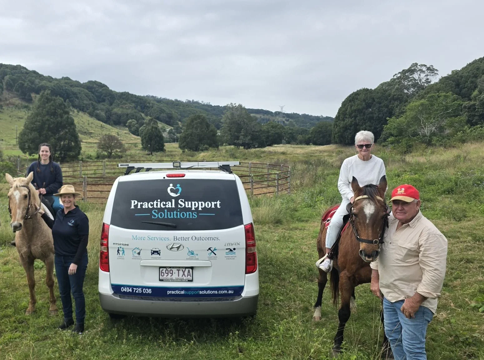 Group of four people with two horses in a grassy field with hills and trees, standing around a white van labeled 'Practical Support Solutions.' Two people are on horseback, while two others stand on the ground.