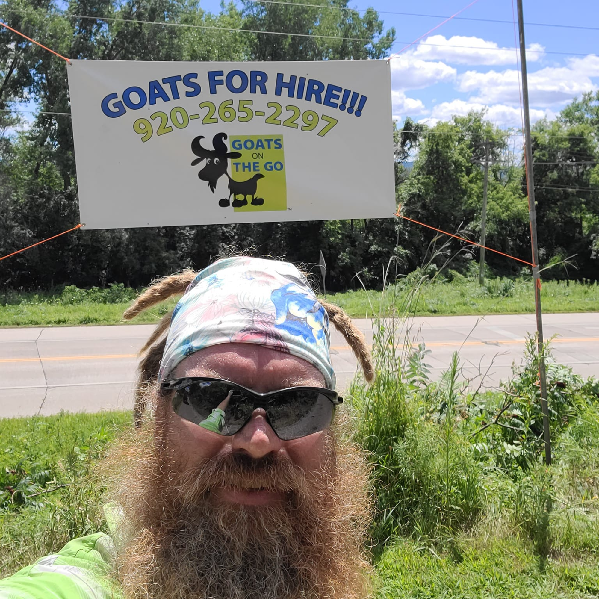 A person with a beard and sunglasses lying on the grass, with their head close to the camera. Behind them, a sign advertising goats for hire, with a cartoon goat and a phone number. The setting is outdoors with trees and a partly cloudy sky.
