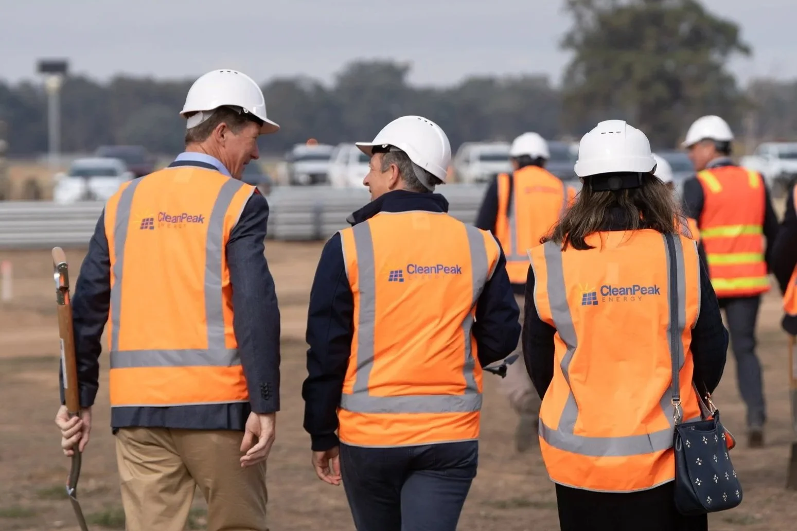 Group of CleanPeak Energy workers in high-vis safety vests and white hard hats walking on a solar farm construction site, with vehicles and trees in the background.