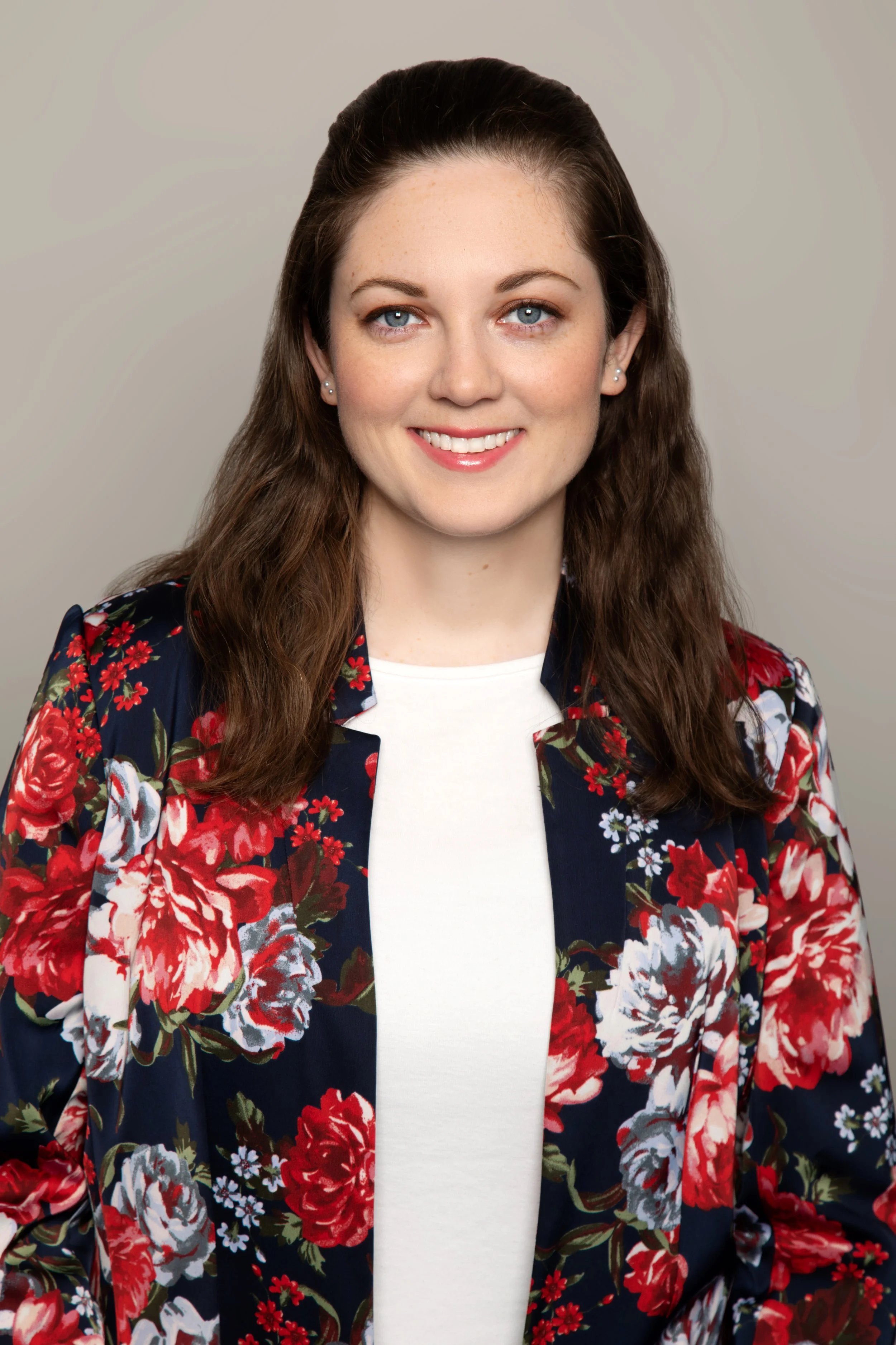 A professional young woman with long brown hair, blue eyes, and light skin, wearing a white top and a navy floral blazer, smiling against a plain gray background.