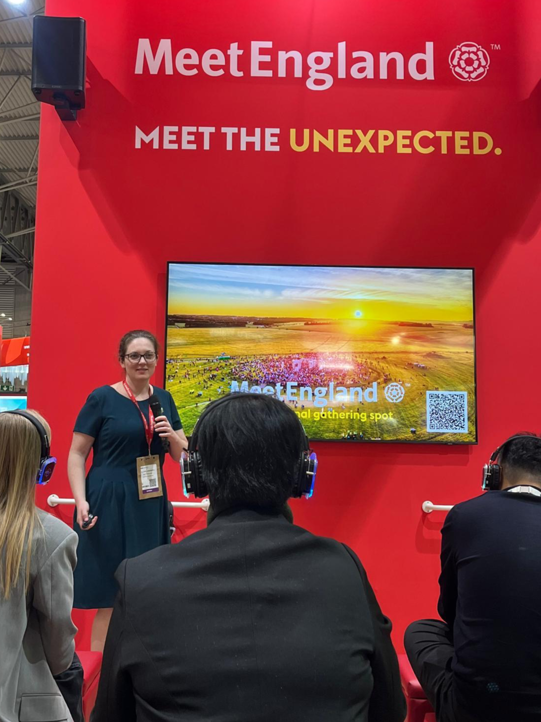 A woman giving a presentation at the MeetEngland booth with a large screen displaying a sunrise over a green field, and people in the audience listening with headphones.