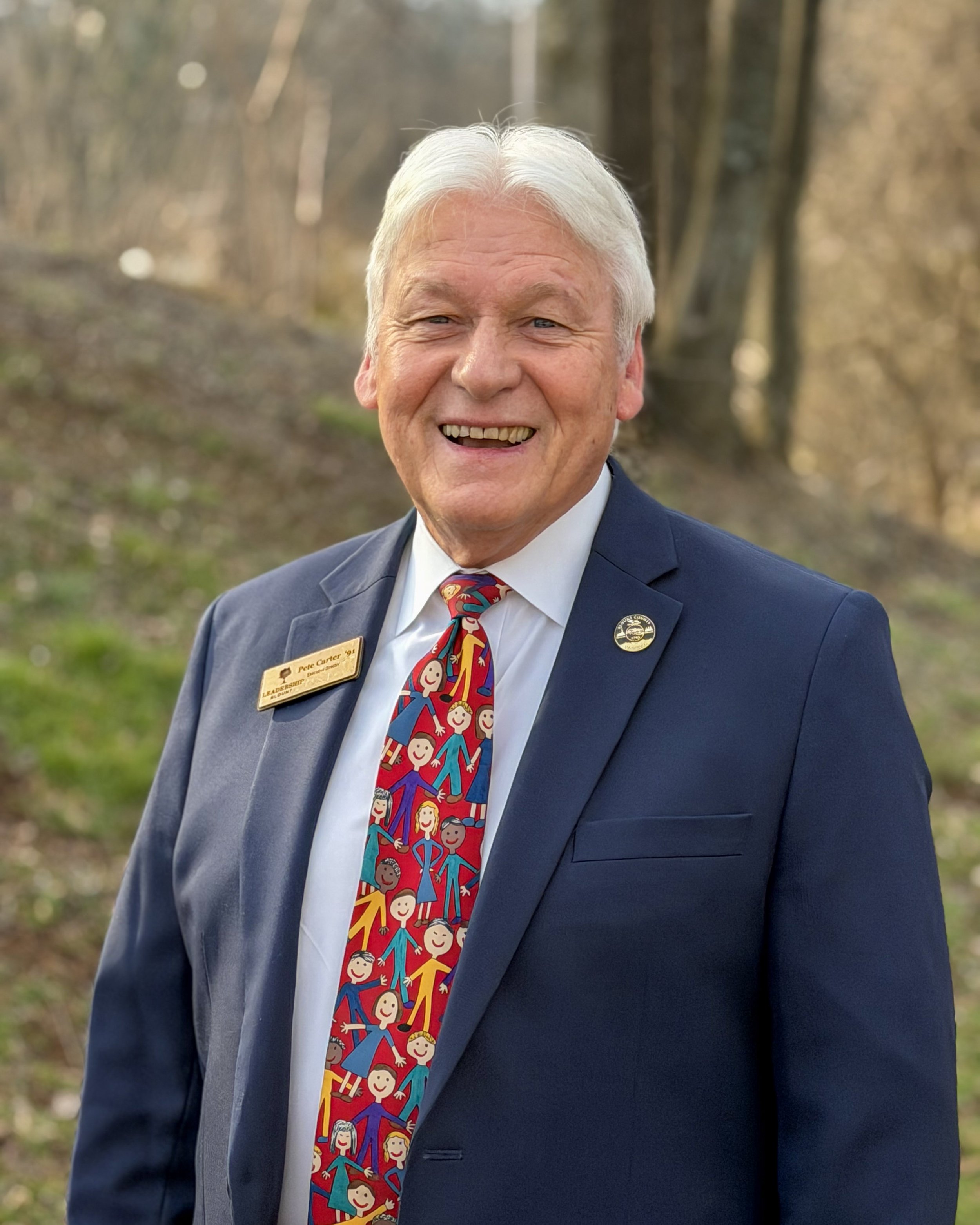 Older man with gray hair smiling in an outdoor setting, wearing a navy suit, a white shirt, and a colorful tie with smiling children's faces, and a gold name tag.