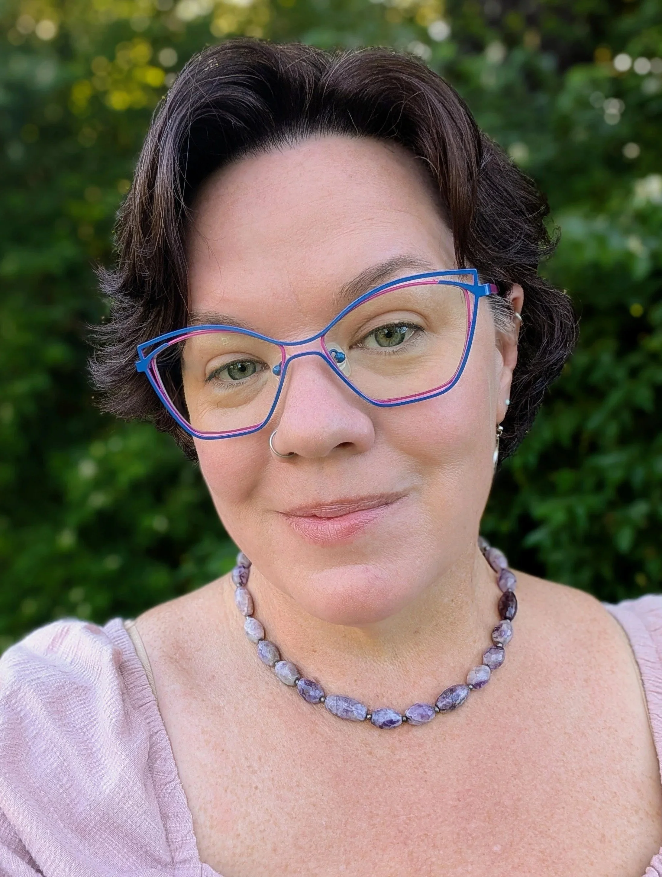 Close-up of a woman wearing purple glasses, a beaded necklace, and a light purple top, with a blurred green outdoor background.