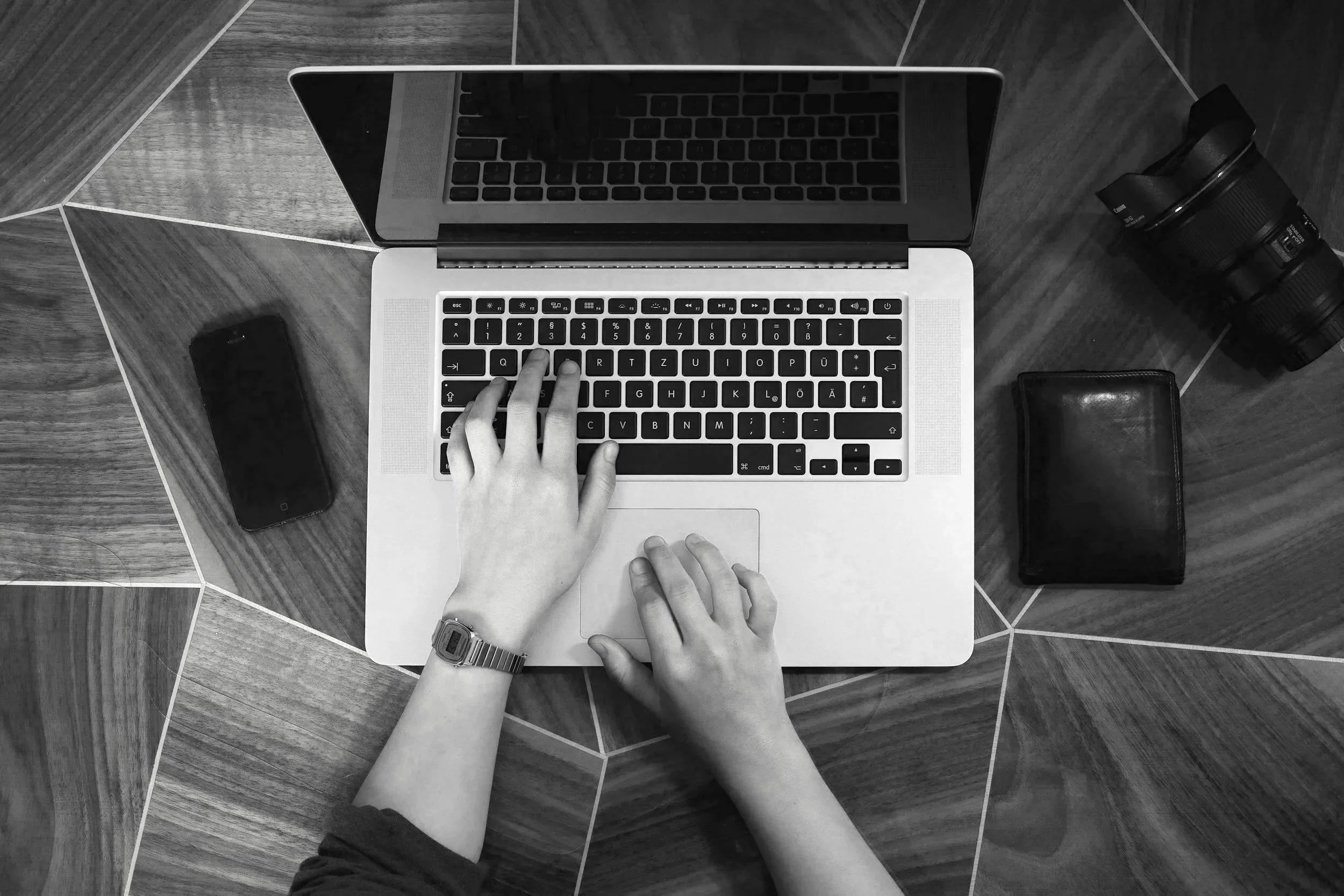 Top down view of a person working on a laptop on a wooden table, with an iPhone, camera lens, wallet, and a wristwatch visible nearby.