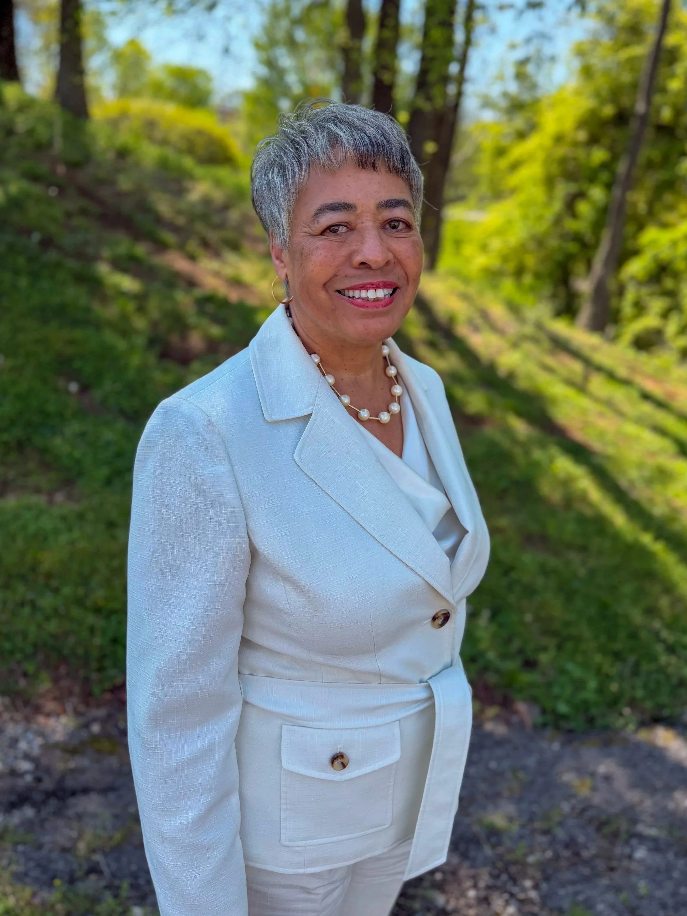 A woman with short gray hair smiling outdoors in a light-colored suit and pearl necklace, standing in front of a green forested area.