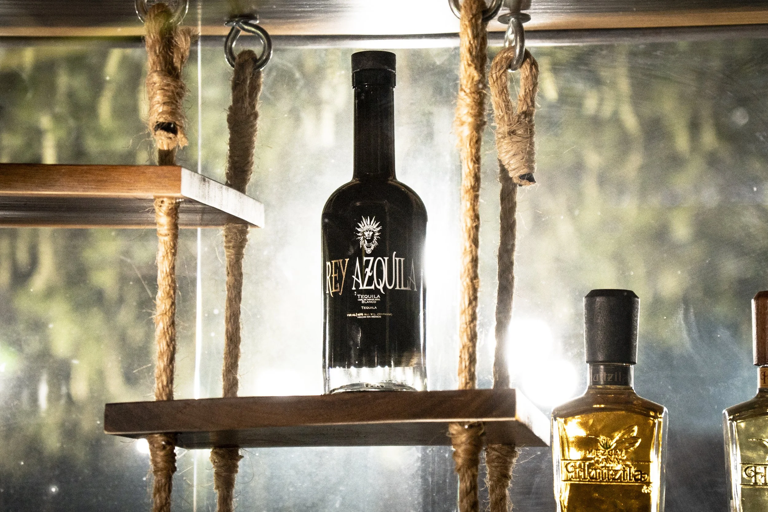A bottle of Rey Azula tequila hangs from a ceiling, flanked by two ropes, with a wooden shelf and other bottles underneath.