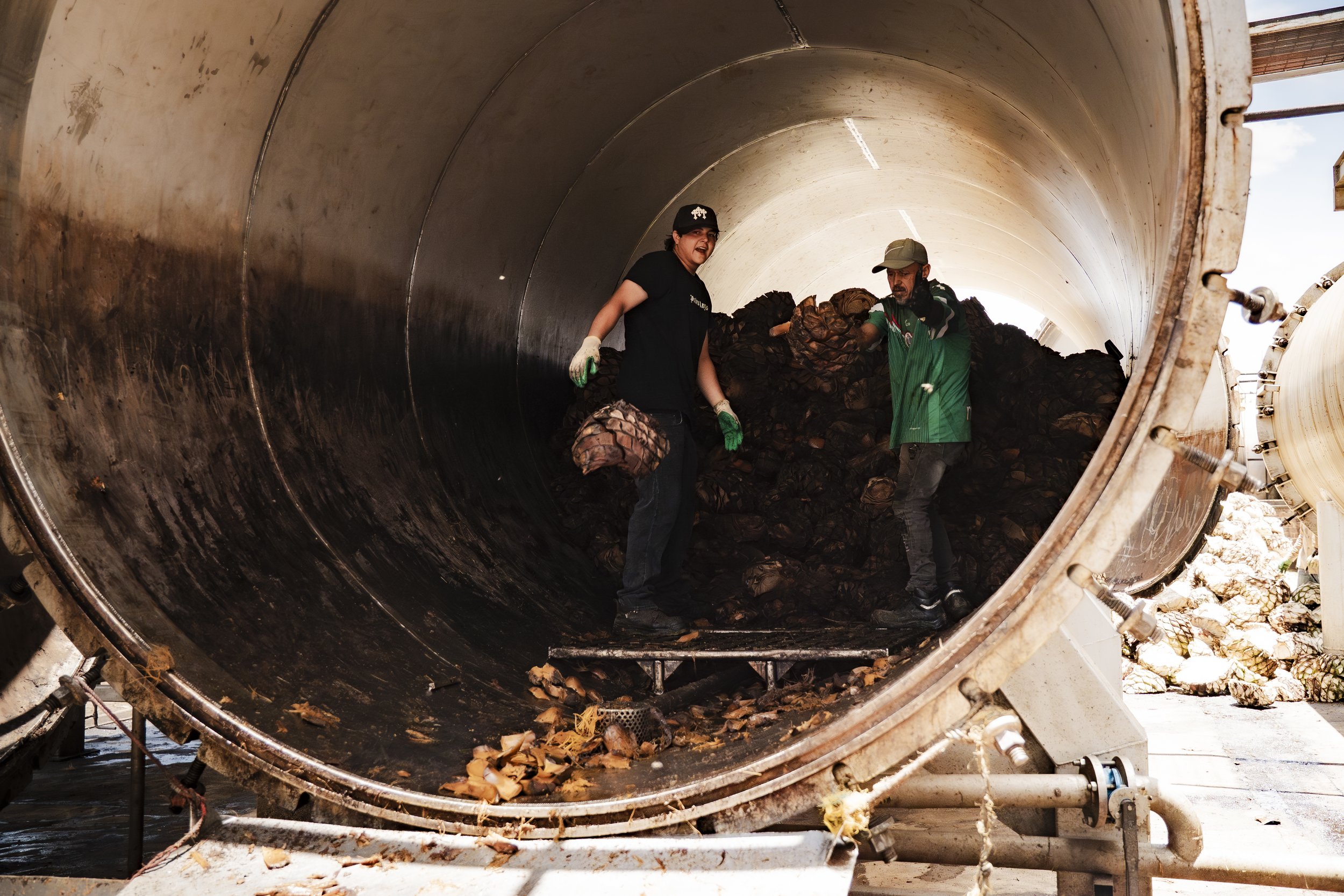 Two workers inside a large industrial metal container or tank, handling a pile of brown organic material, possibly shells or husks, with sunlight visible from the outside.