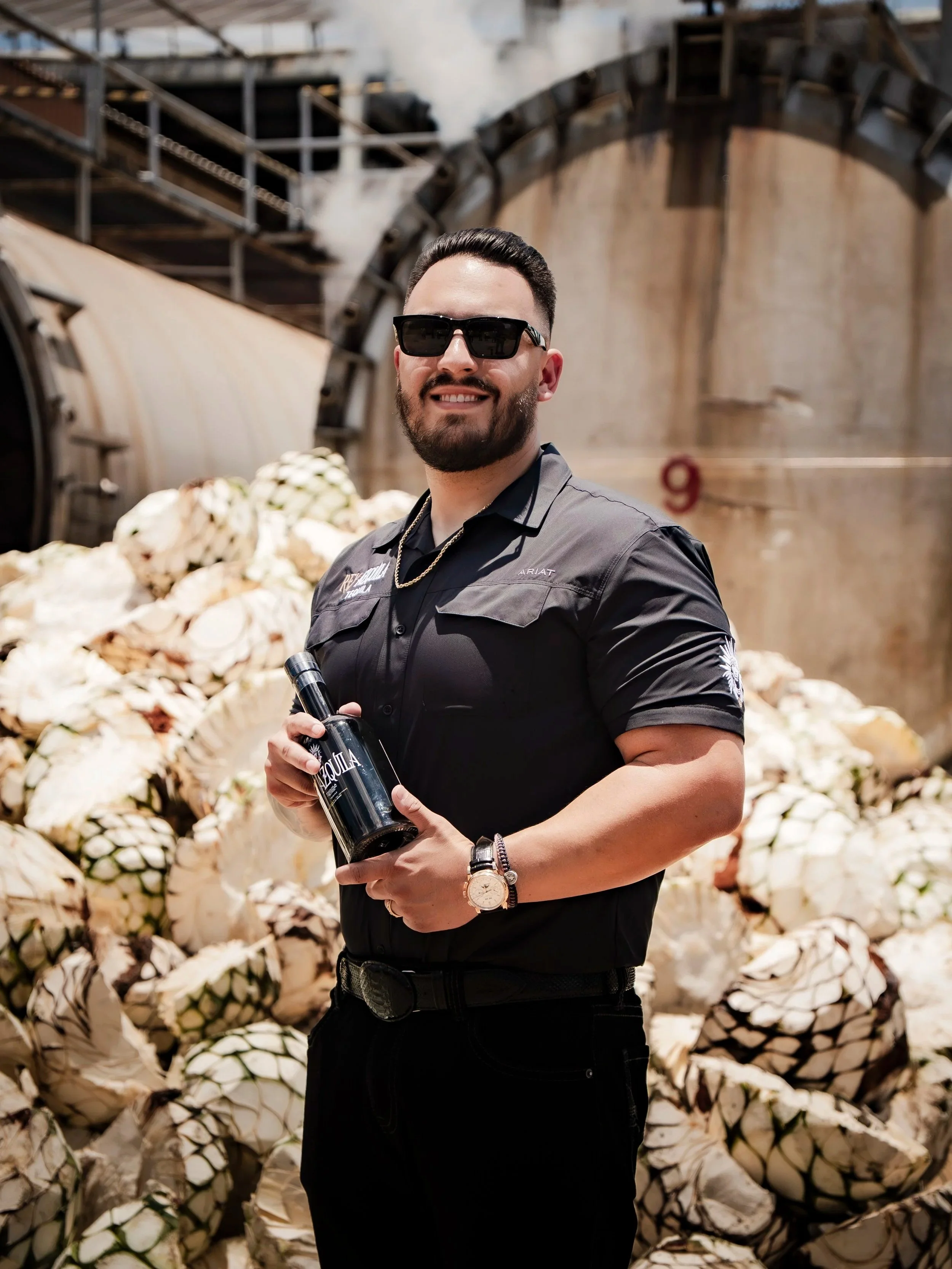 A man with a beard and sunglasses smiling, holding a bottle in front of a large pile of artichokes, in an industrial setting with steam and metal structures in the background.