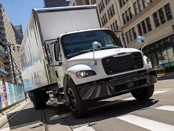 A white delivery truck turning right on a city street with tall buildings on both sides.