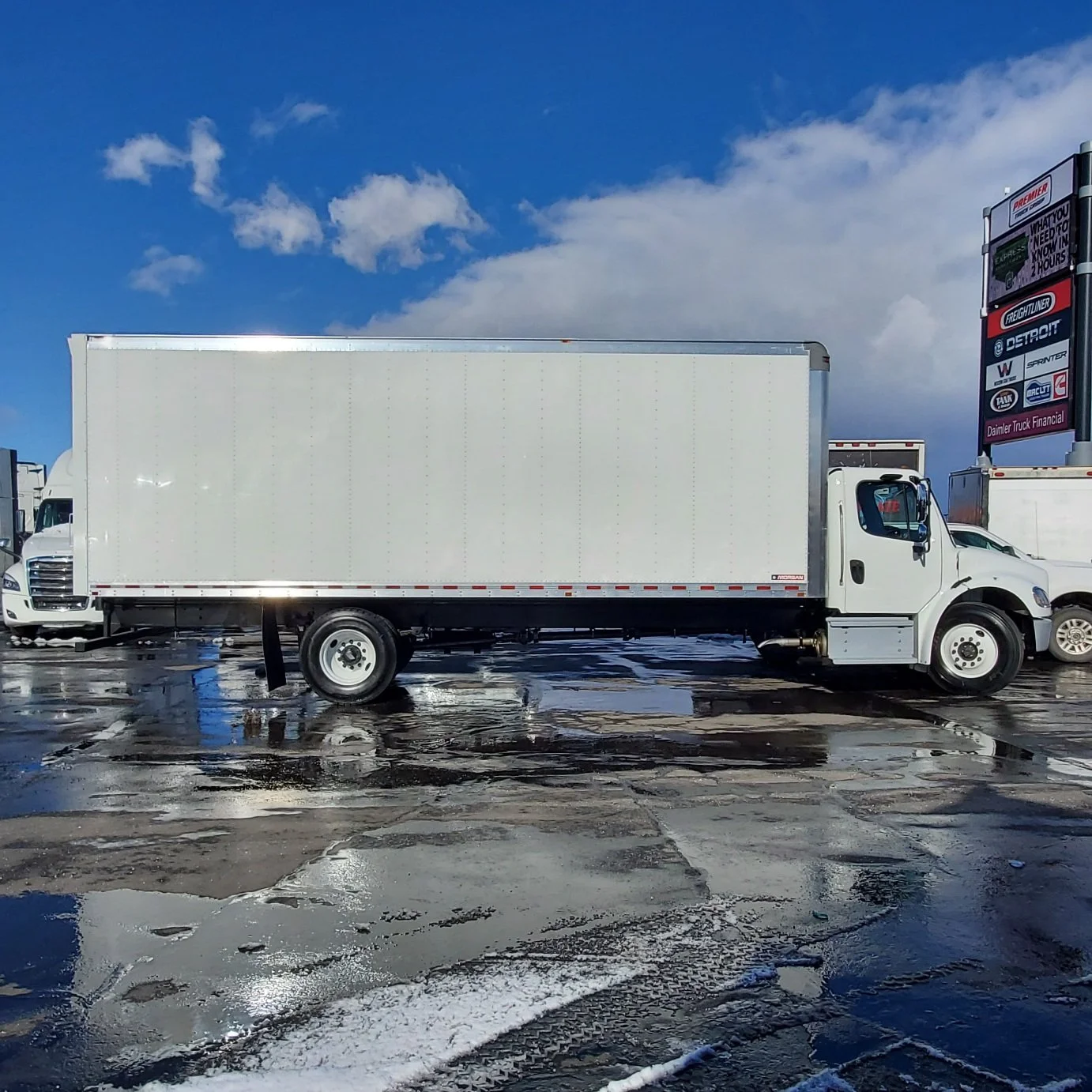 White cargo truck parked on a wet, icy lot with puddles and patches of snow, under a partly cloudy blue sky, with signage for trucking companies in the background.