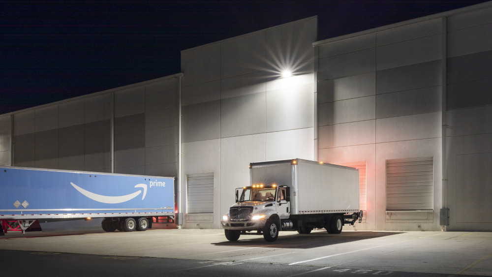 A warehouse loading dock at night with an Amazon Prime delivery truck and a semi-truck parked outside.