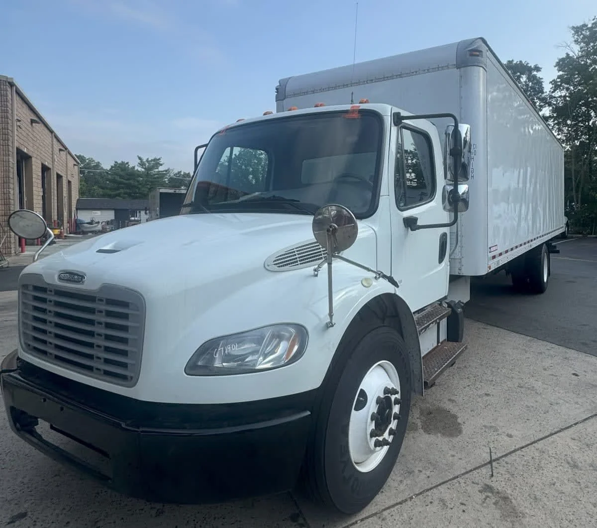 White box truck parked on pavement in front of a building, with side mirrors and tires visible.