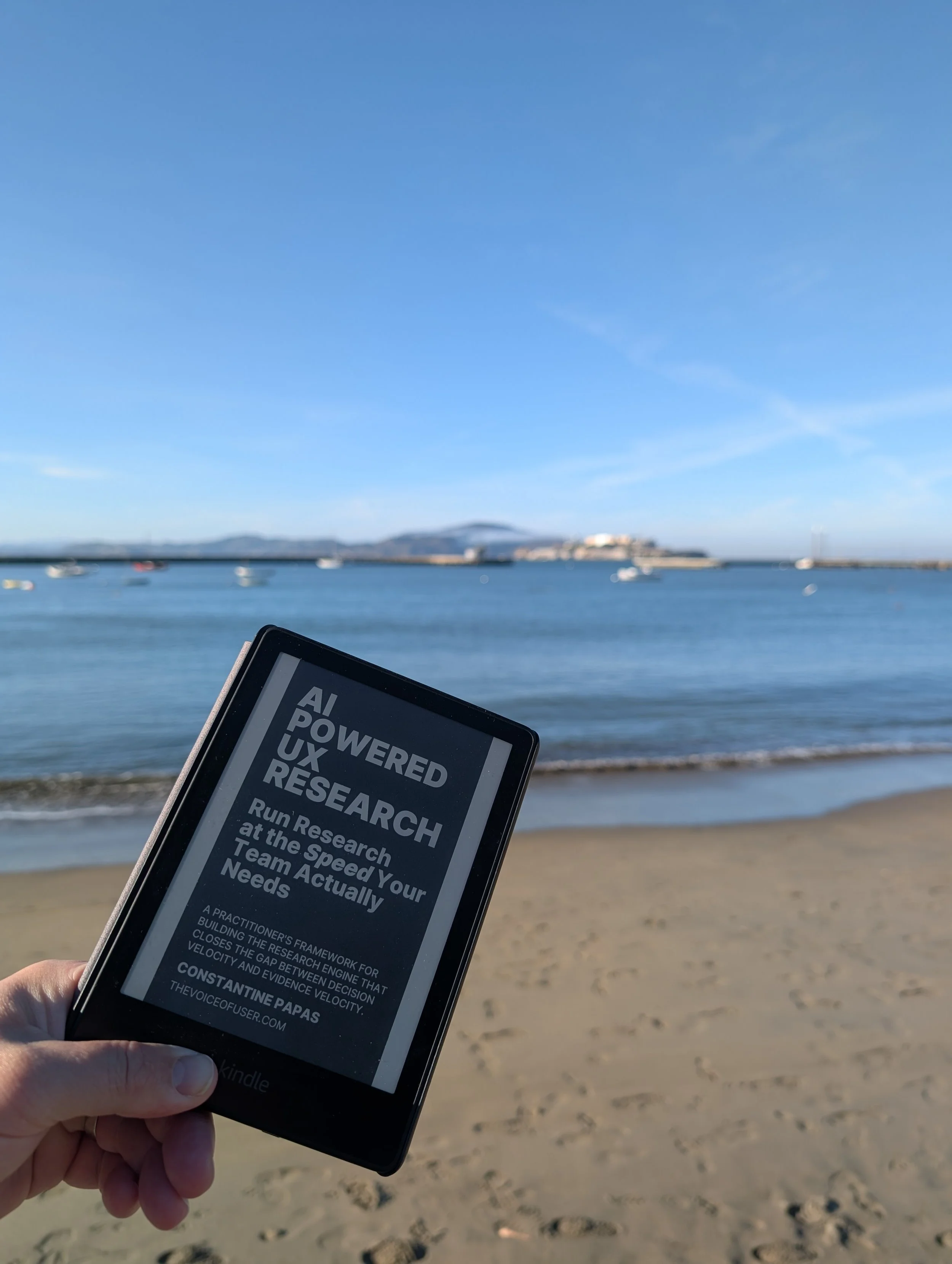 A Kindle with a book on the screen about how to run UX Research at speed without sacrificing rigor or quality in the new AI era on the beach at Aquatic Park near Fort Mason the day before the conference.