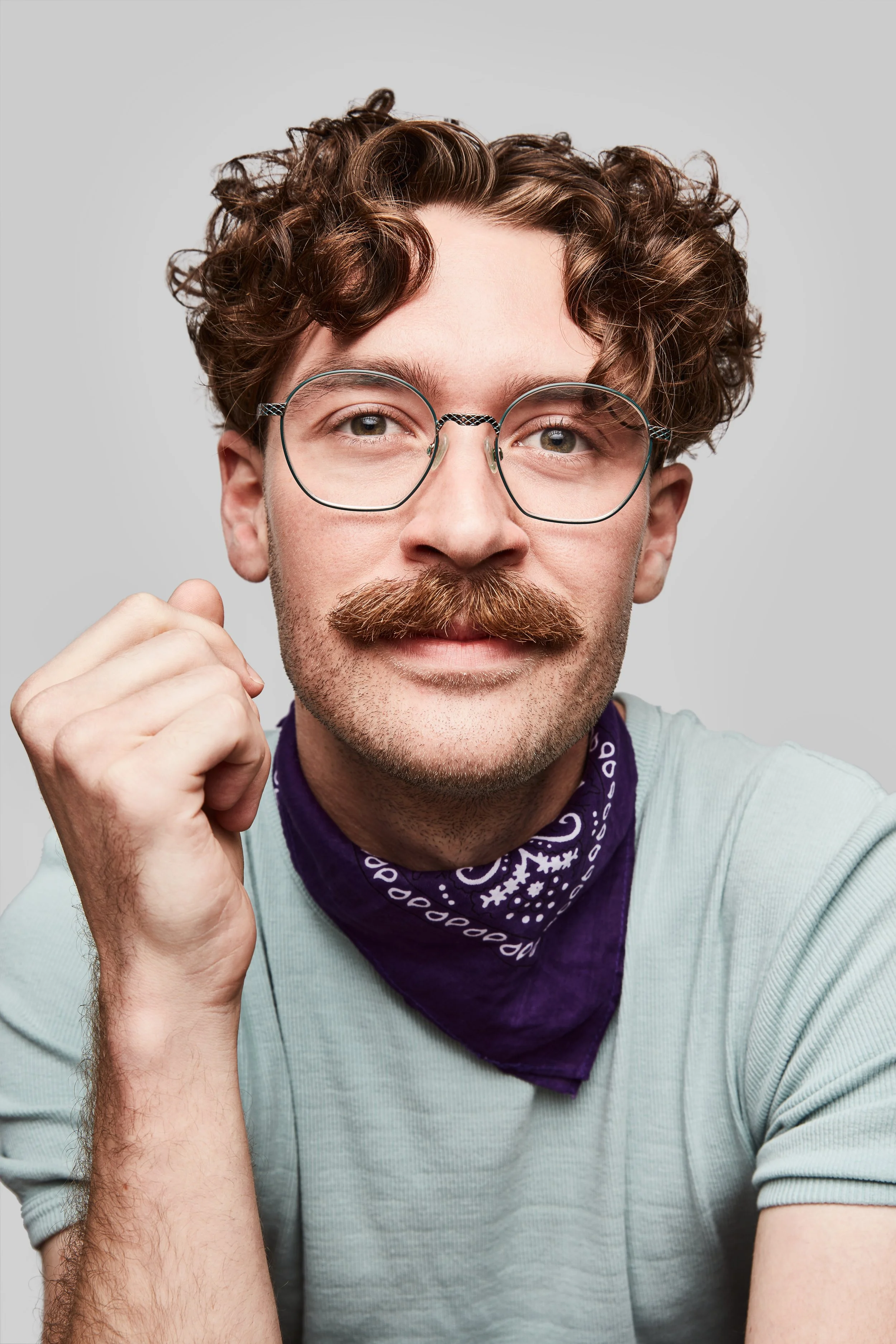 A young man with wavy brown hair, glasses, and a mustache, wearing a light blue shirt and purple bandana, poses with his hand near his face against a plain gray background.