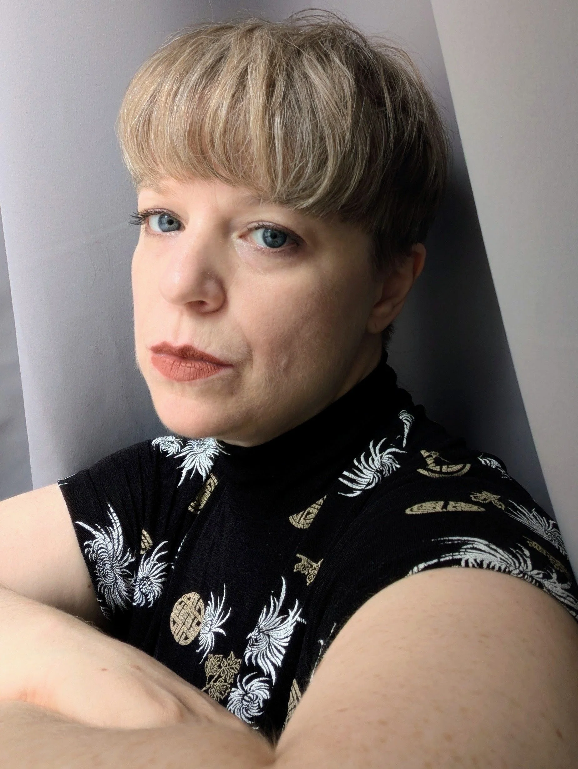 Breanne Oryschak. A woman with short blonde hair and blue eyes, wearing a black shirt with white and gold floral patterns, is taking a selfie in front of a plain gray wall.
