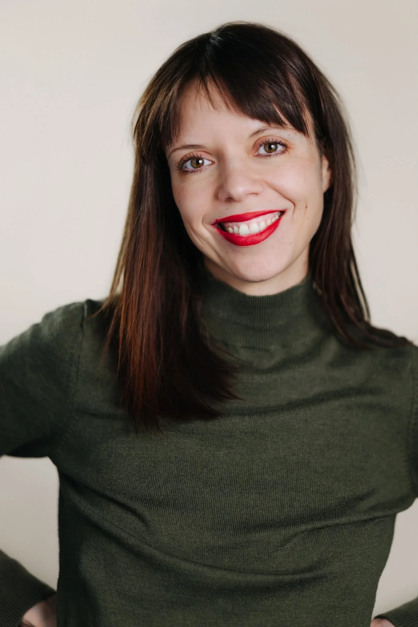 Theresa Stork. A woman with ginger hair and fair skin is smiling and wearing red lipstick, a green turtleneck sweater, and is standing against a plain light-colored background.