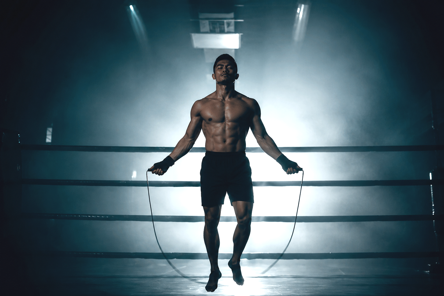 A shirtless male athlete with a muscular physique and athletic build jumping rope in a boxing ring, with a bright light behind him and surrounded by fog or smoke.
