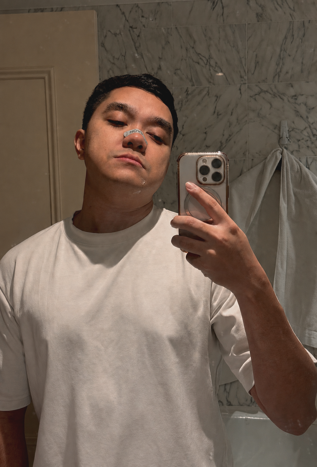 A young man taking a selfie in a bathroom mirror, wearing a white t-shirt, with a small bandage on his nose and chin, and a marble wall background.