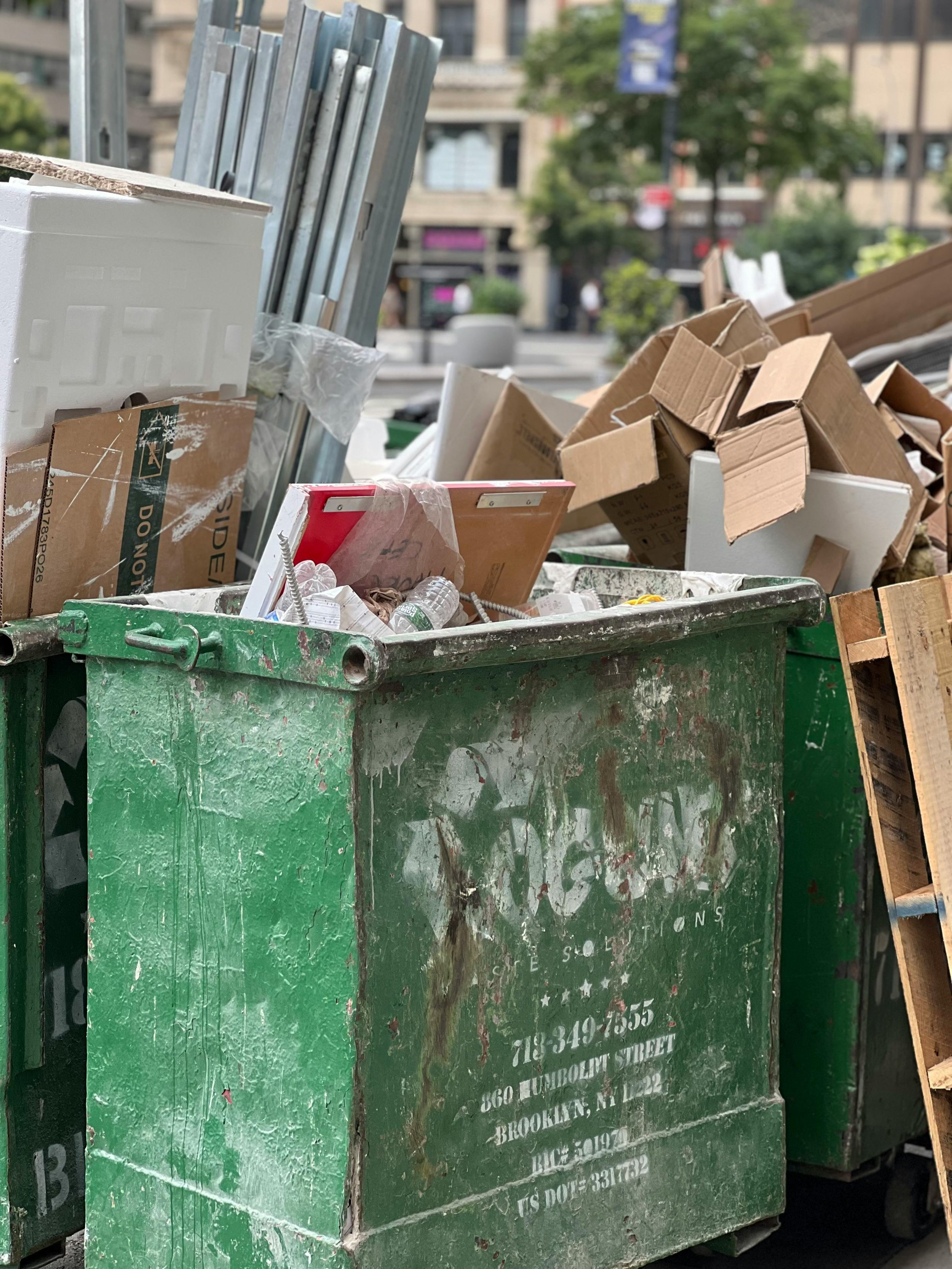 Green dumpster overflowing with cardboard, paper, and small trash, with city buildings and trees in the blurred background.