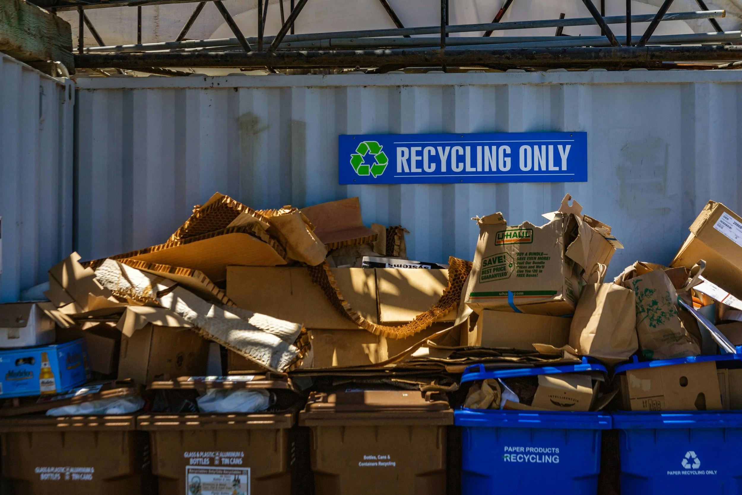 A blue recycling bin labeled 'RECYCLING ONLY' containing crumpled cardboard and paper waste. There are other recycling bins below, labeled for different recyclable materials, with items like cardboard, paper, and bottles visible inside.