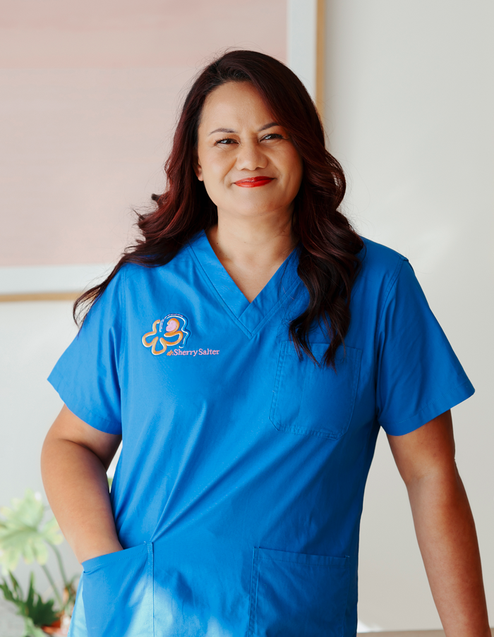A woman wearing blue medical scrubs with a name embroidered on the chest walks confidently, smiling at the camera in a bright room.