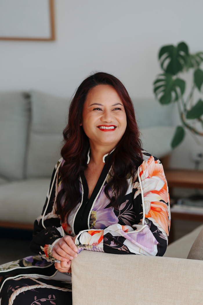 A woman with long dark hair and red lipstick sitting on a beige couch in a living room, smiling and wearing a colorful floral-patterned jacket.