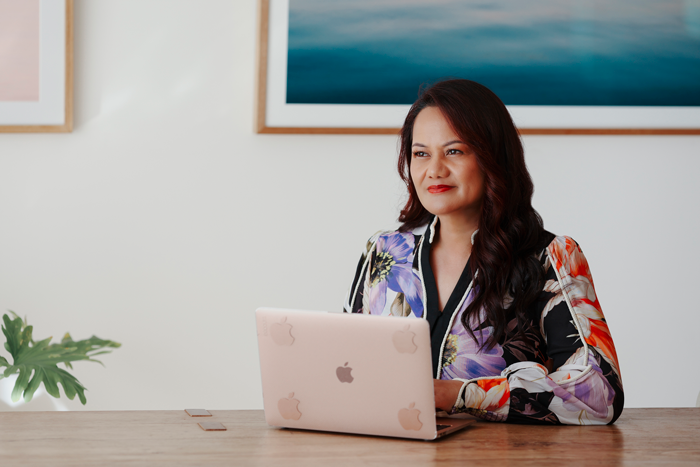 A woman with long dark hair sits at a wooden desk with a laptop, looks slightly to the side with a thoughtful expression, and is in a room with framed artwork on the wall and a green plant.