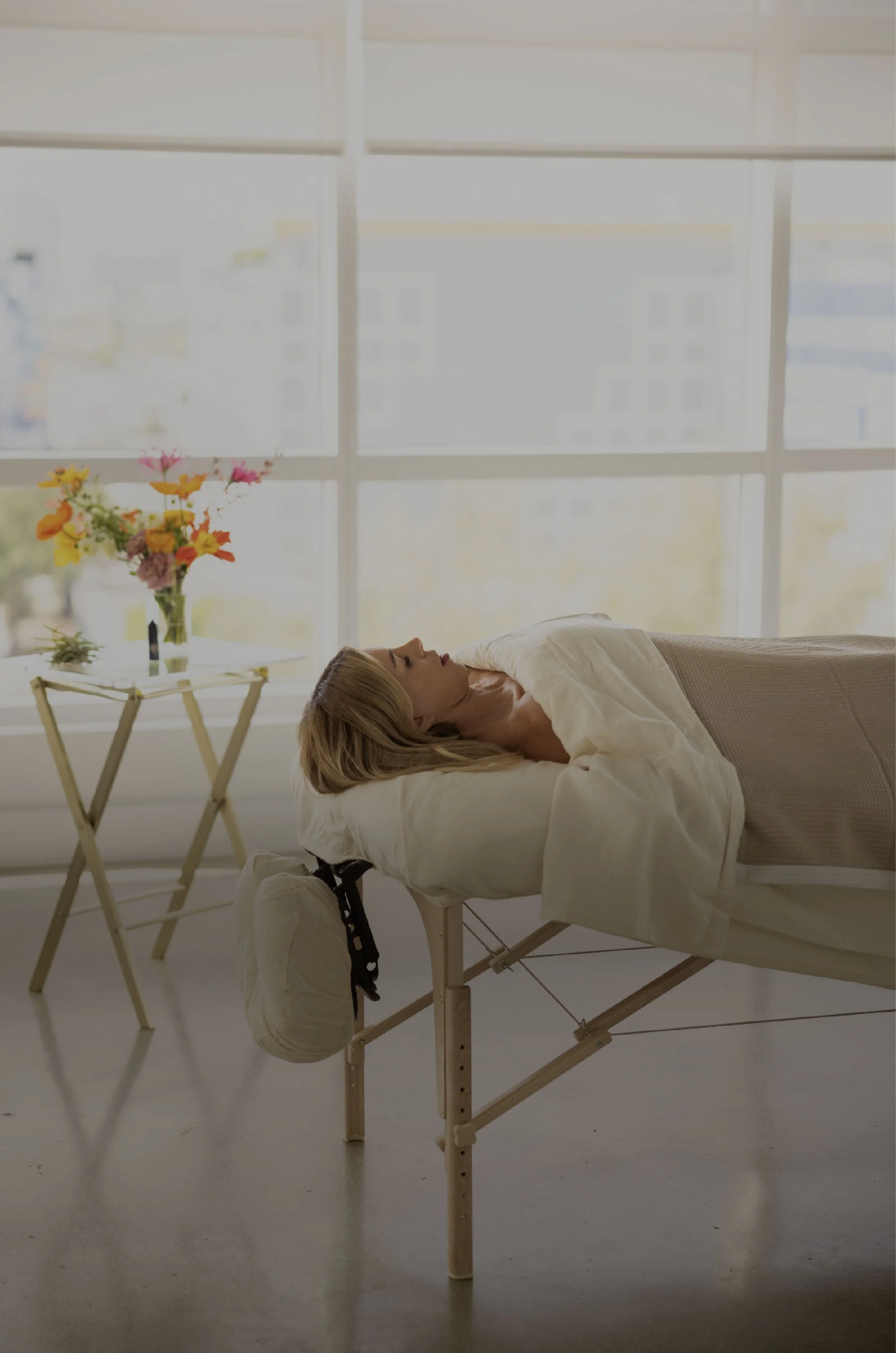 A woman lying on a massage table in a bright room with large windows, a small table with flowers, and a pair of sneakers hanging from the corner of the table.