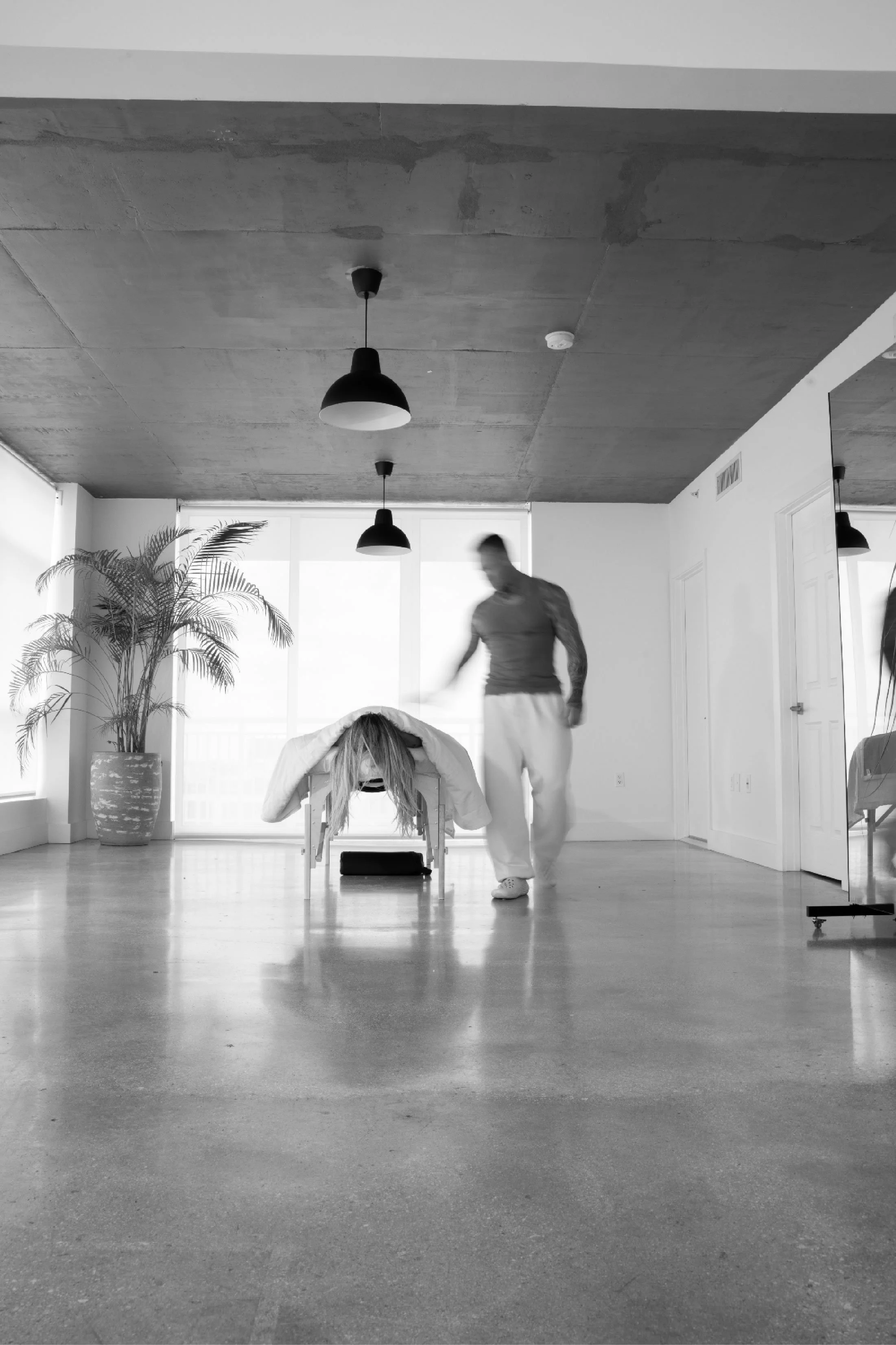 A woman with long hair bending over and leaning forward on a lounge chair in a spacious room, with a man walking past in the background. The room has large windows, a potted plant, and pendant lights hanging from a concrete ceiling.