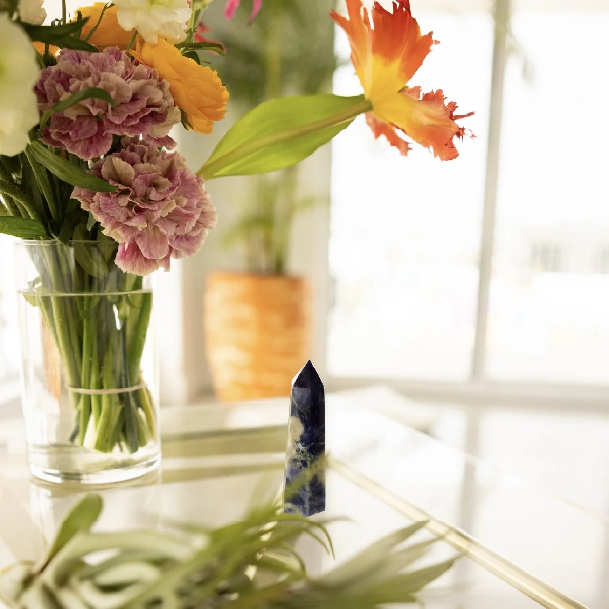Close-up of a bouquet of colorful flowers in a glass vase, with a blue and white crystal standing on a white surface in the foreground. Bright natural light streams in from a window in the background.