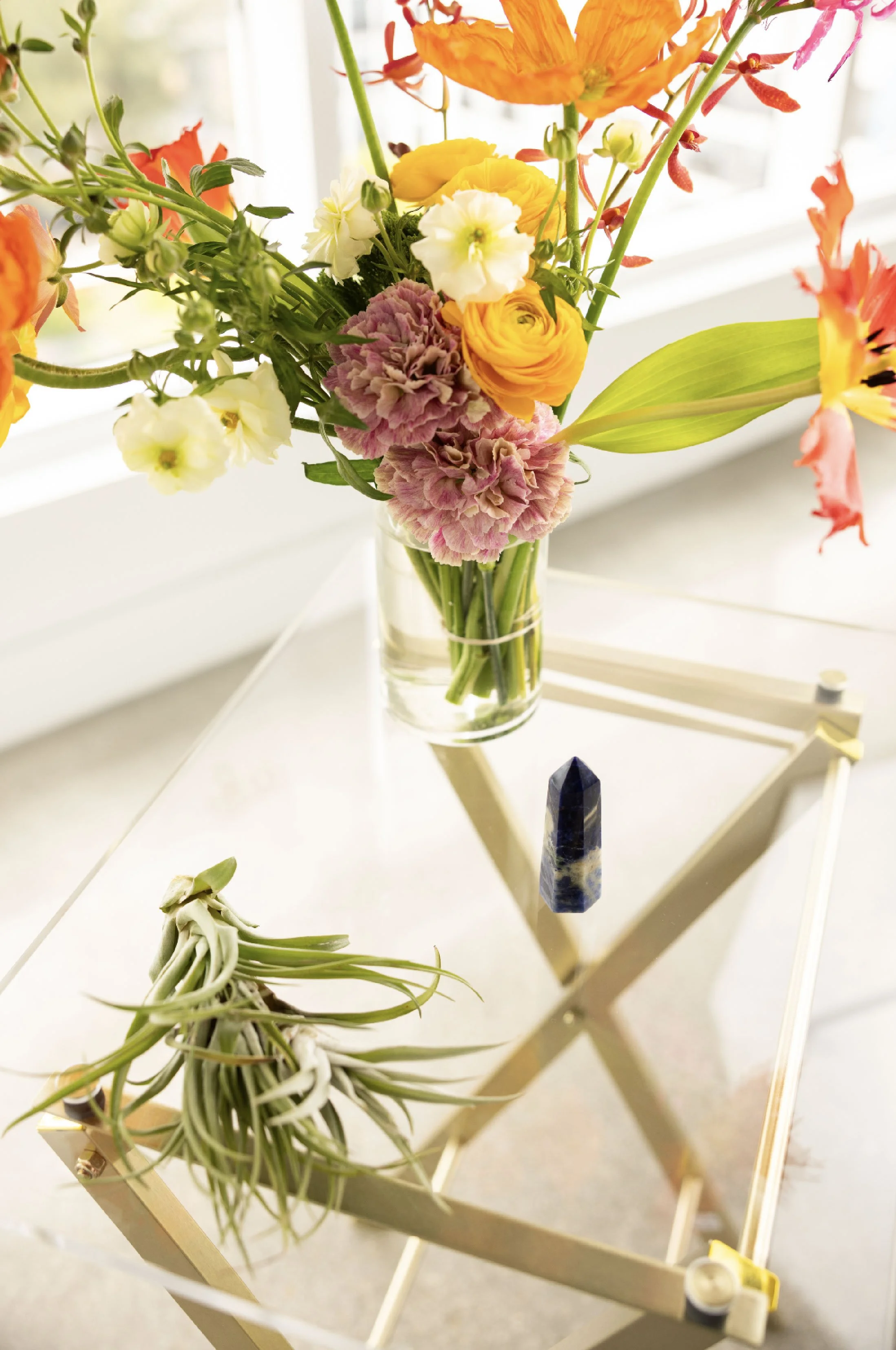 A bouquet of mixed flowers including orange, yellow, pink, and white blooms in a glass vase on a white surface near a window, with a few air plants and a pole with a dark stone on the table.