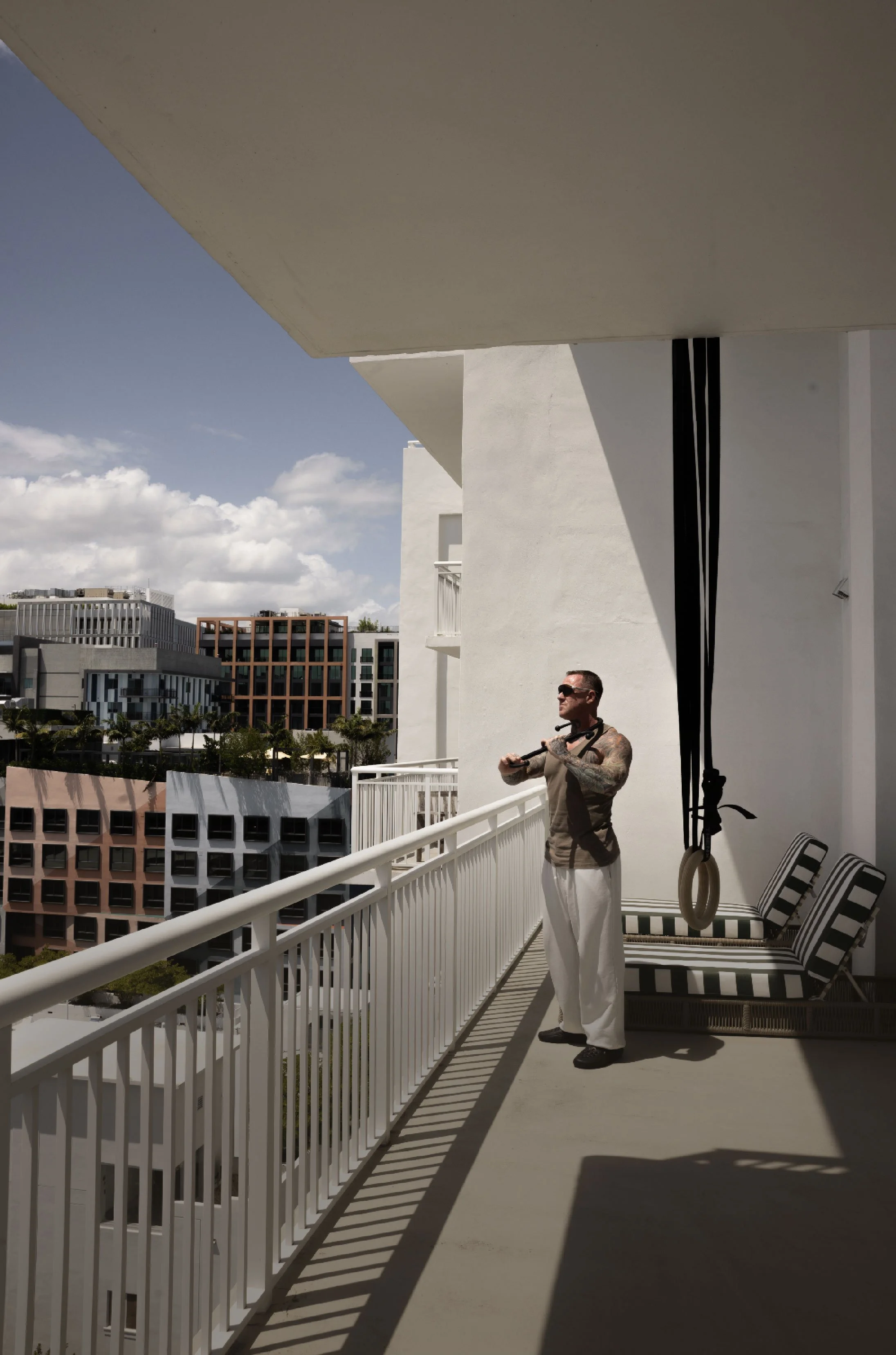 A man holding a resistance band on a balcony with white railings and striped outdoor furniture. Shadows cast by the balcony railing create striped patterns on the floor. Modern buildings and a blue sky with scattered clouds are visible in the background.