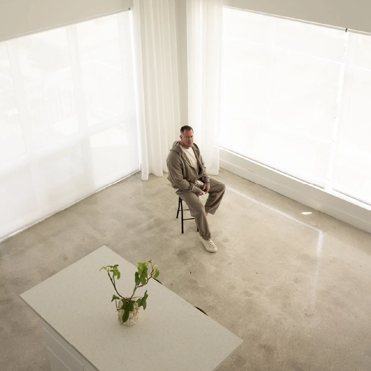 A man sitting on a stool in a bright white room with large windows and white curtains, facing forward. A white table with a potted plant is in the foreground.