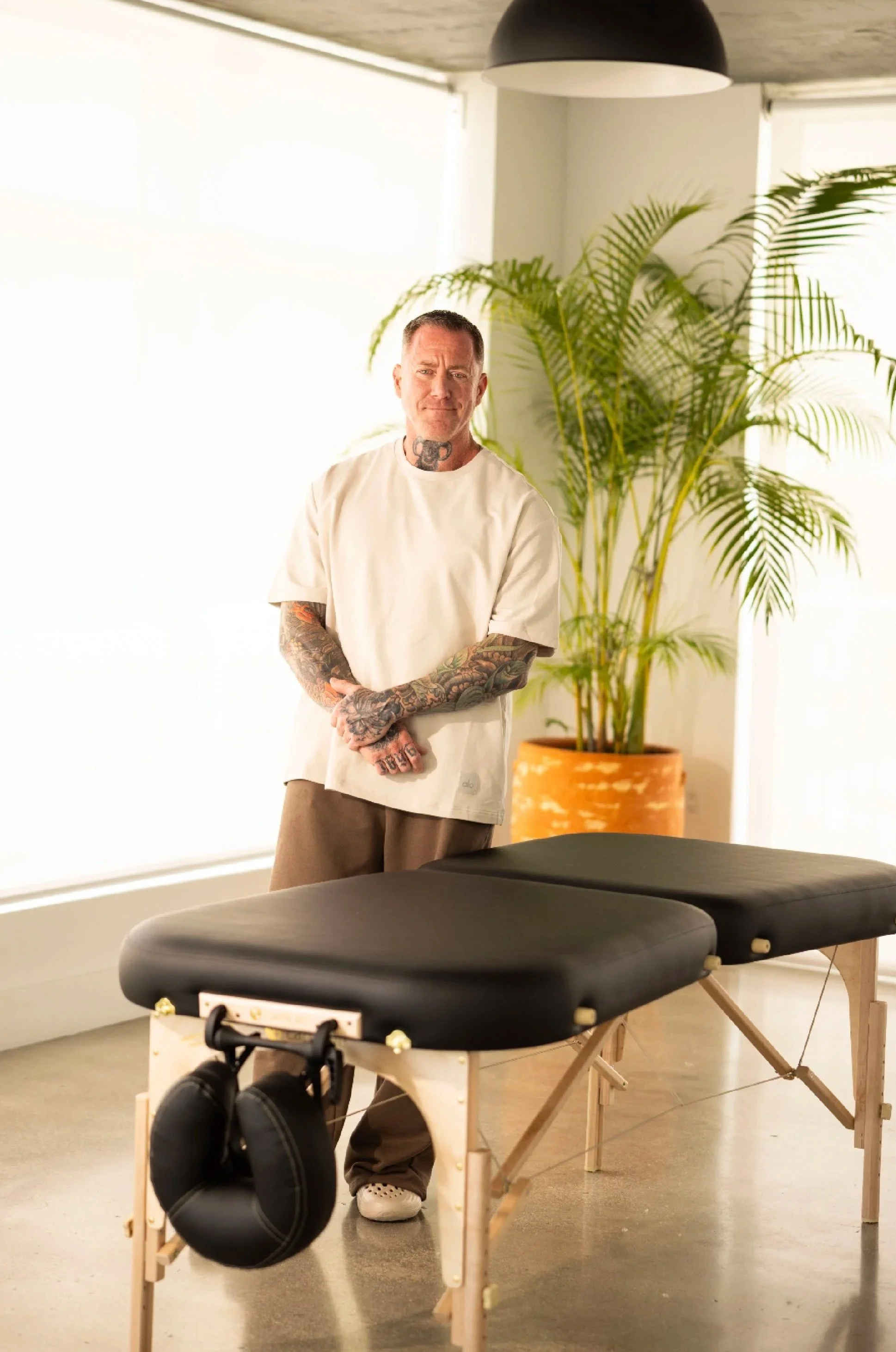 A man with tattoos on his arms and neck standing in a room with a large potted plant behind him, near a window. In the foreground, there is an empty massage or chiropractic table with a pair of headphones hanging from one corner.