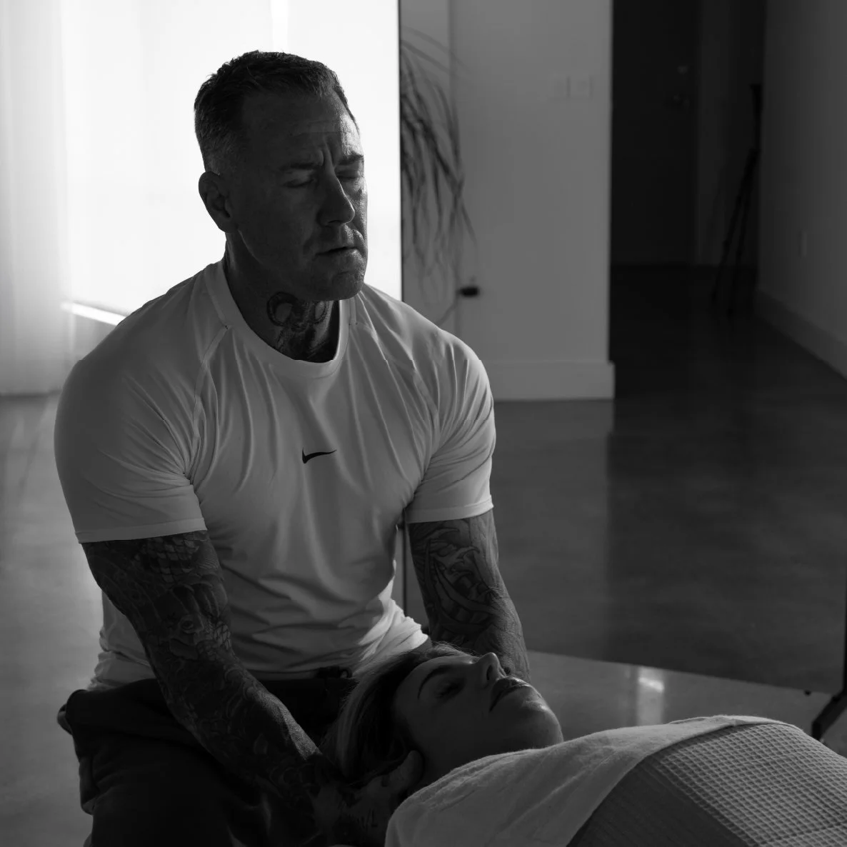A man with tattoos is giving a massage or treatment to a woman lying on a bed in a dimly lit room.