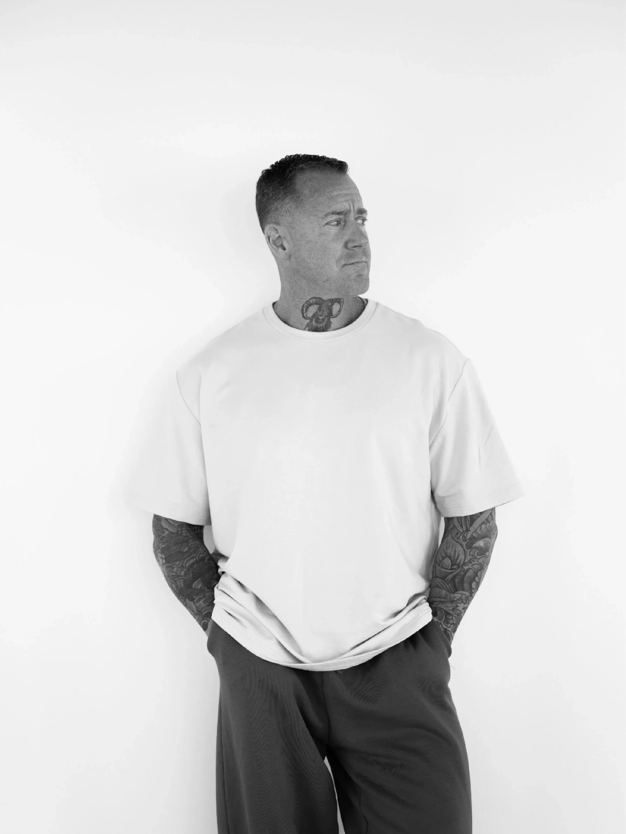 Black and white photo of a man with tattoos on his arms and neck, wearing a white t-shirt and black pants, standing against a plain white background, looking to the side.