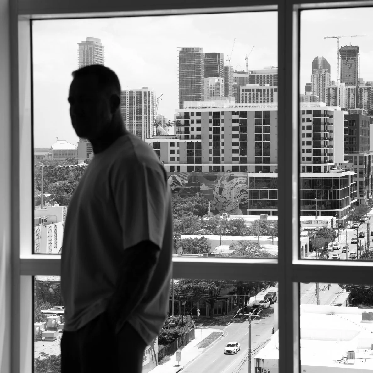 Silhouette of a man standing by a window overlooking a cityscape with tall buildings and cranes.