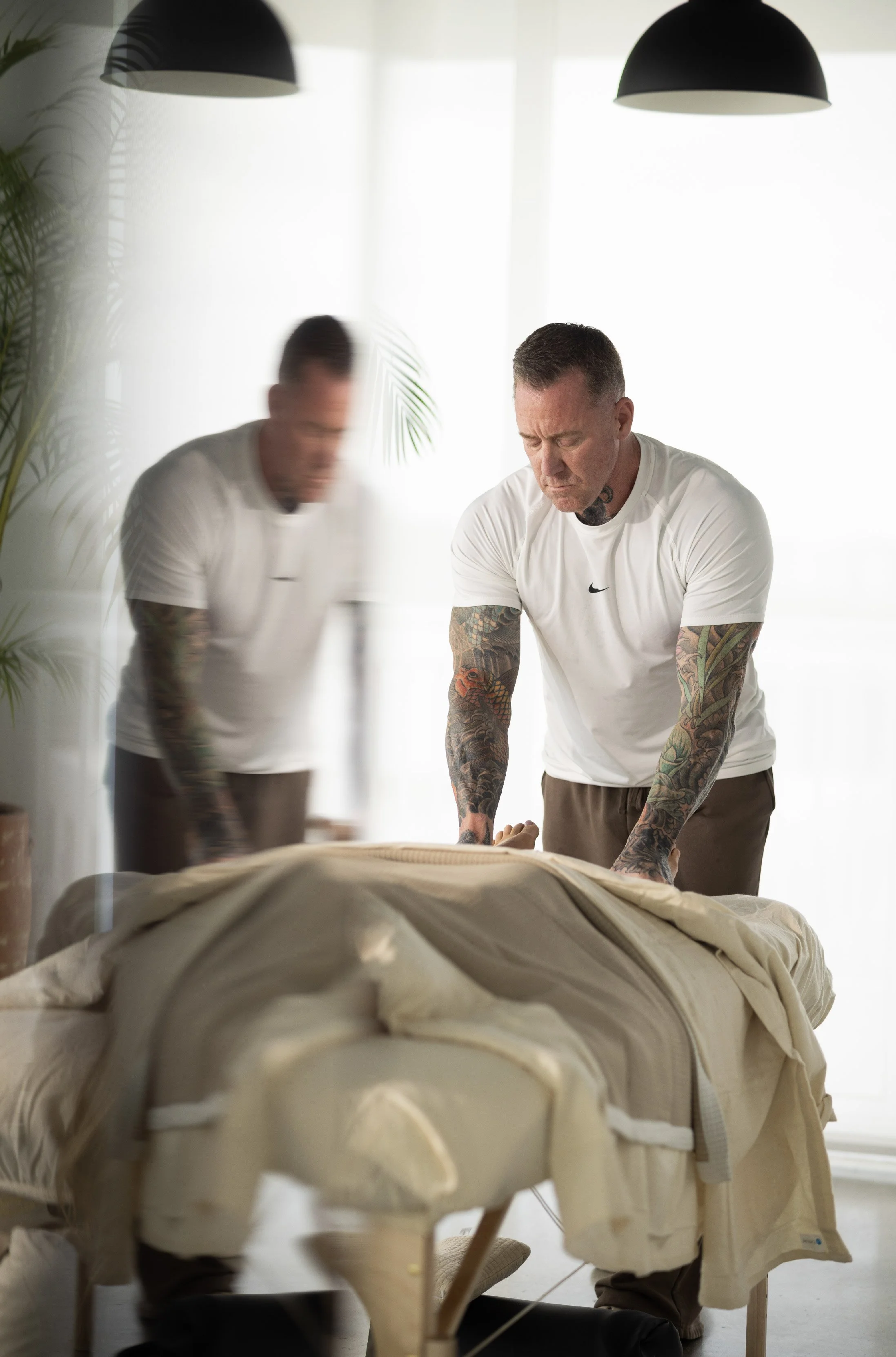 Man performing a massage or physical therapy on a person lying on a treatment table in a bright, modern room with large windows and black hanging lights.