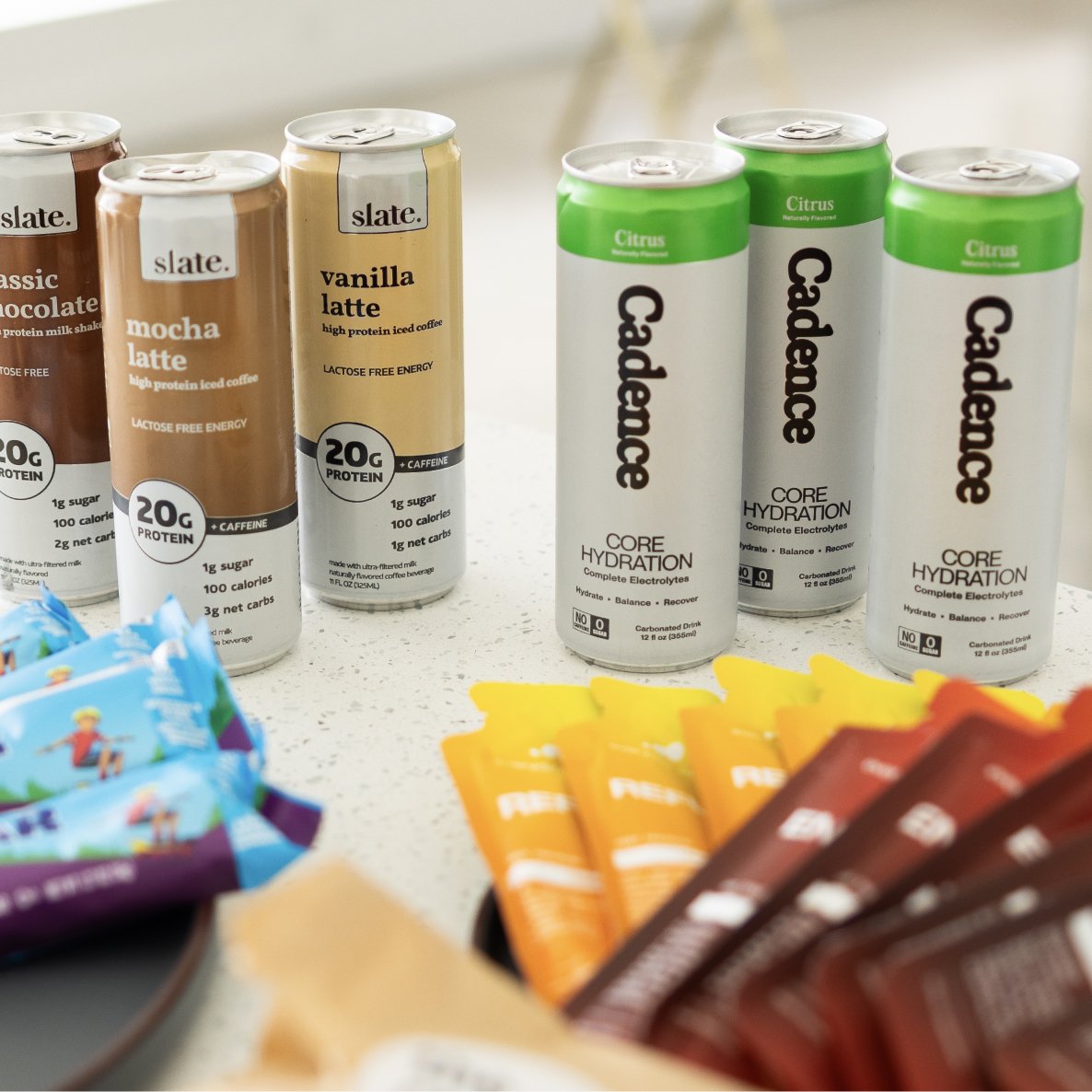 Canned beverages on a counter, including Slate mocha and vanilla latte drinks, and Caladence core hydration electrolyte drinks, with colorful snack wrappers in the foreground.