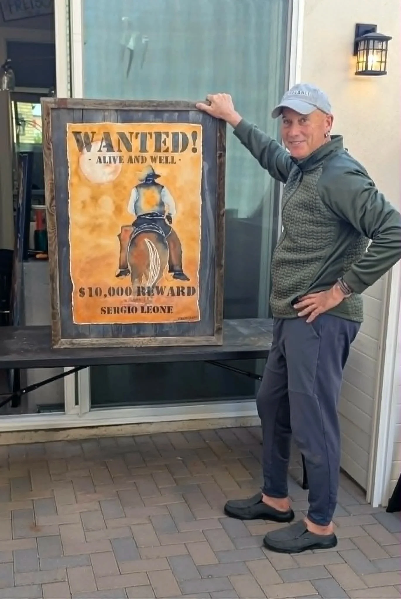 A man with short hair, wearing a gray baseball cap, dark gray jacket, and gray shoes, standing next to a wanted poster outside a building. The poster shows a person riding a horse and offers a $10,000 reward for Sergio Leone.