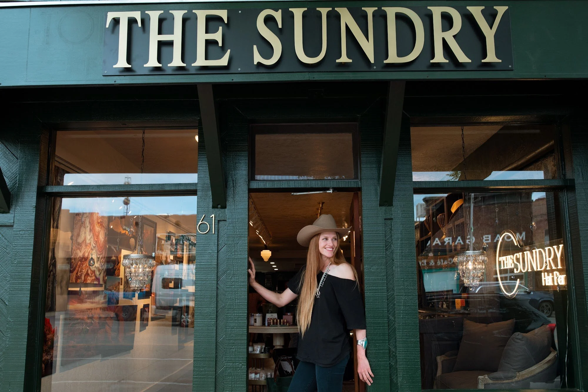 A woman with long red hair wearing a cowboy hat and black top is standing in the doorway of a store called 'The Sundry.' She is smiling and looking to her right, holding the door frame with her left hand. The store's interior and a reflection of cars on the street are visible through the glass.