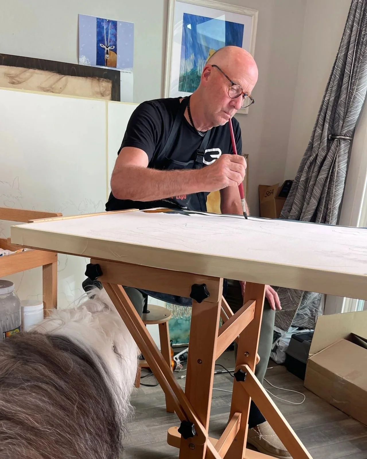 Peter painting on a large canvas set up on an easel in a room with artwork hanging on the wall, a curtain, and a dog in the foreground.