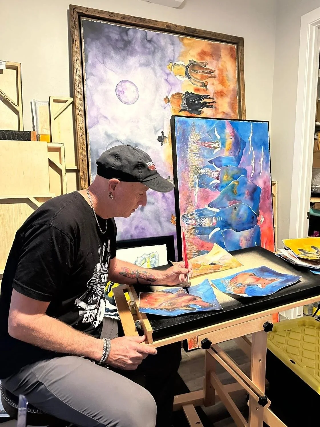 Peter sitting at a work table, painting colorful water-themed animal art with elephants and aquatic life, surrounded by completed paintings and framed artwork on the walls.