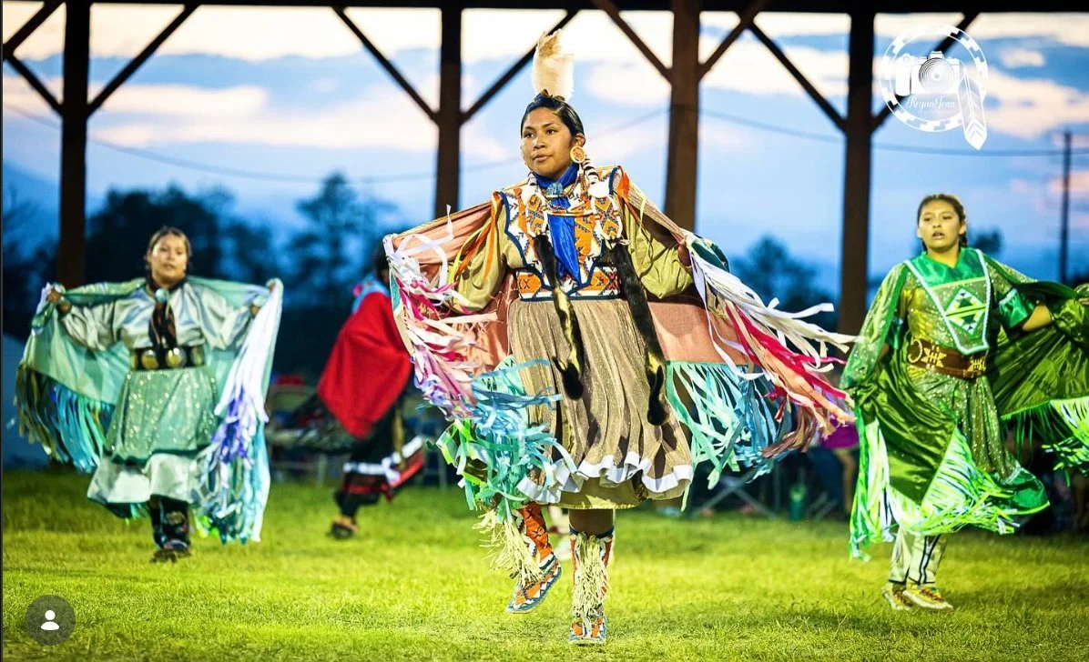 Native American women in traditional colorful attire dancing outdoors during a cultural celebration at sunset.