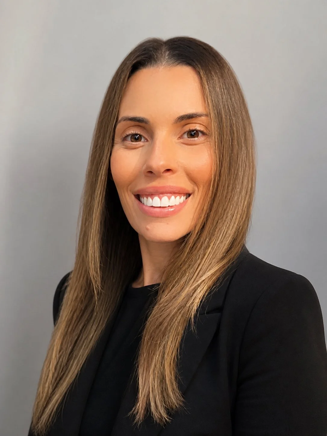 A woman with long light brown hair, smiling, wearing a black blazer, against a plain grey background.