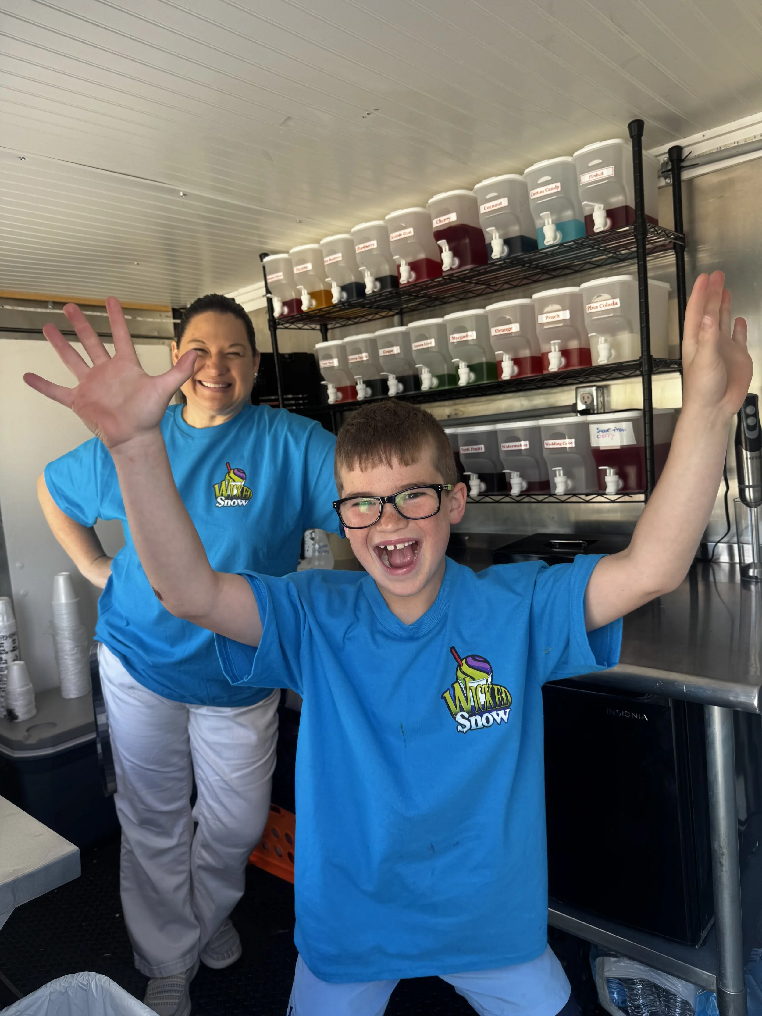 A woman and a young boy, both wearing bright blue shirts with 'Wicked Snow' logos, are smiling and waving at the camera inside a food stand with colorful beverage dispensers in the background.