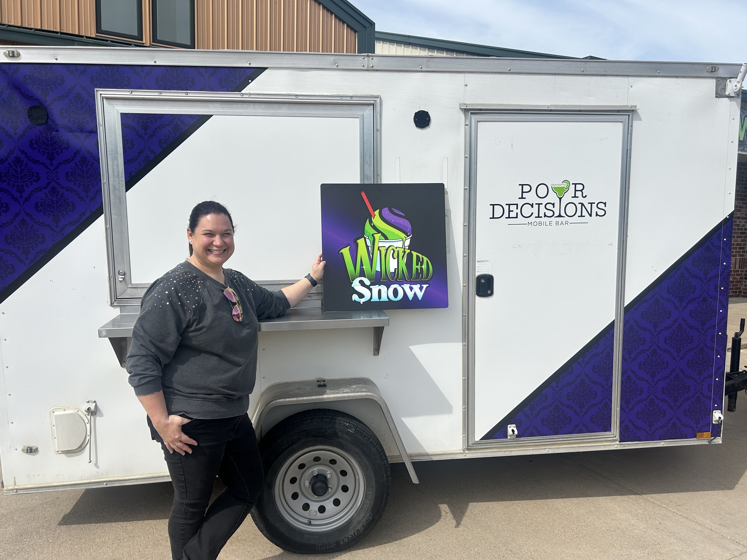 A woman smiling and holding a sign that says "Wicked Snow" stands next to a white mobile bar trailer with the branding "Pour Decisions" on the side. The trailer has decorative purple and black design elements.