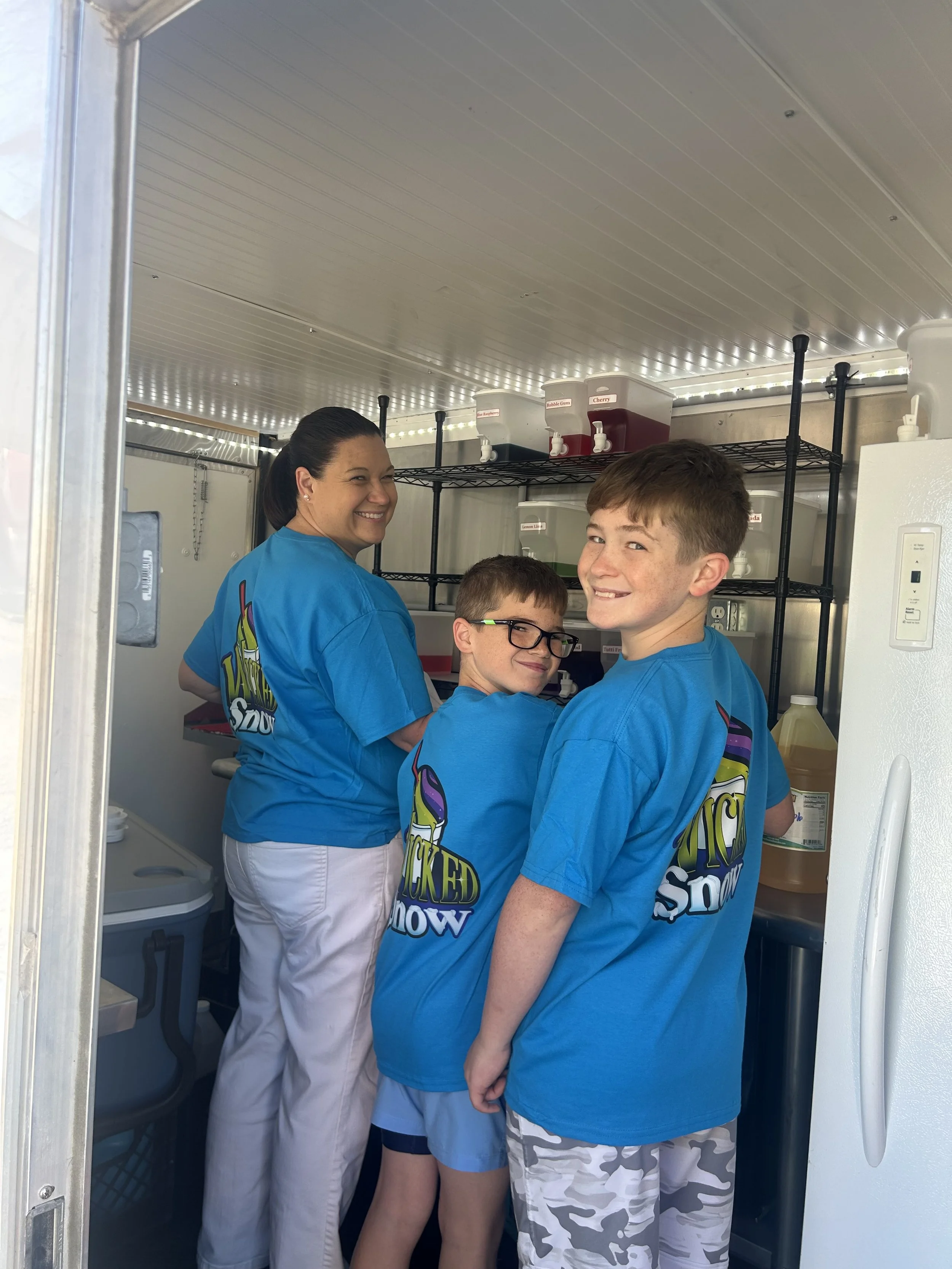 A woman and three boys inside a small kitchen, all wearing matching blue T-shirts, smiling, standing close together, with shelves of syrup bottles labeled with flavors in the background.