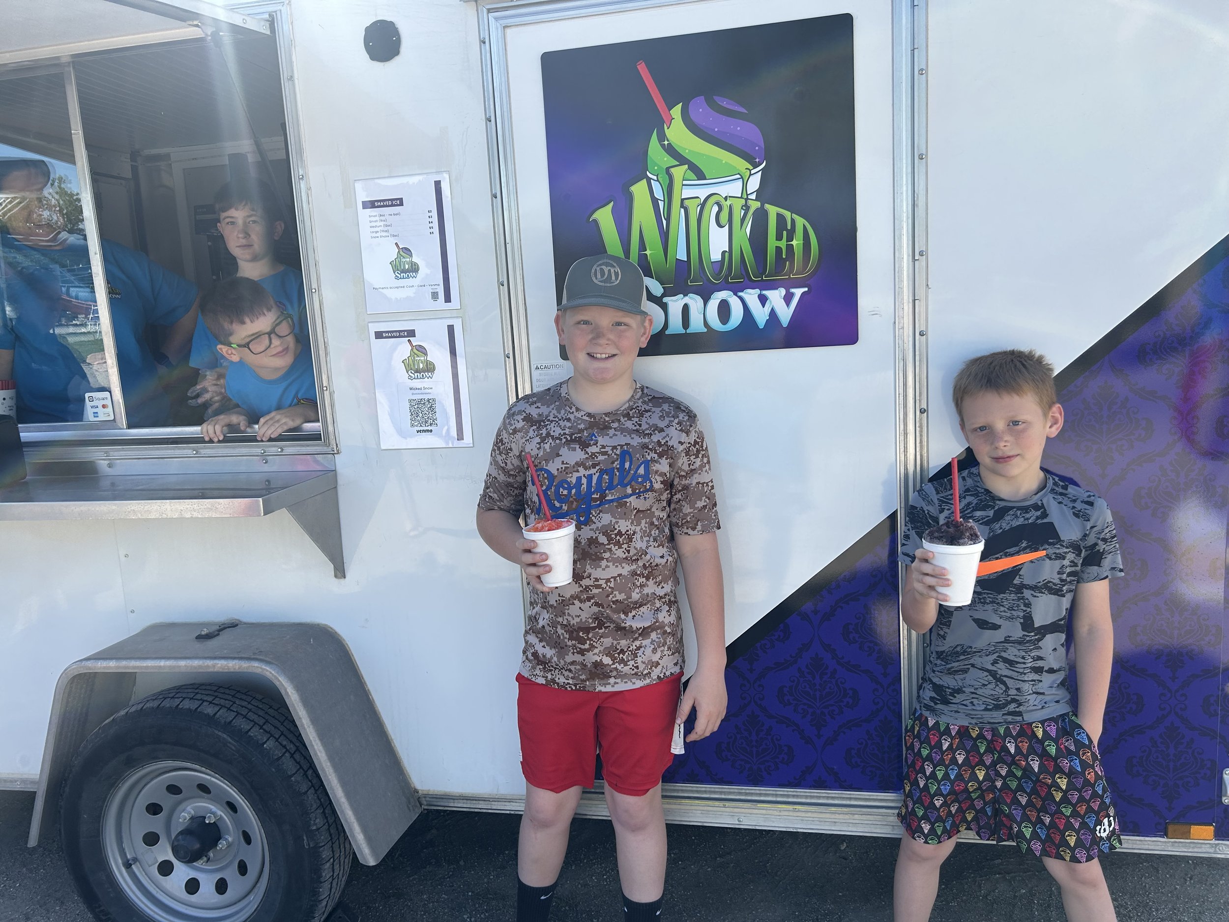 Three boys standing in front of a frozen treat truck. Two of the boys are holding cups of shaved ice with red and purple syrup and straws. The truck has a large colorful sign with a snow cone and the words 'Wicked Snow'. The boys are smiling and wearing casual clothing, with one boy wearing a cap and camouflage shirt.
