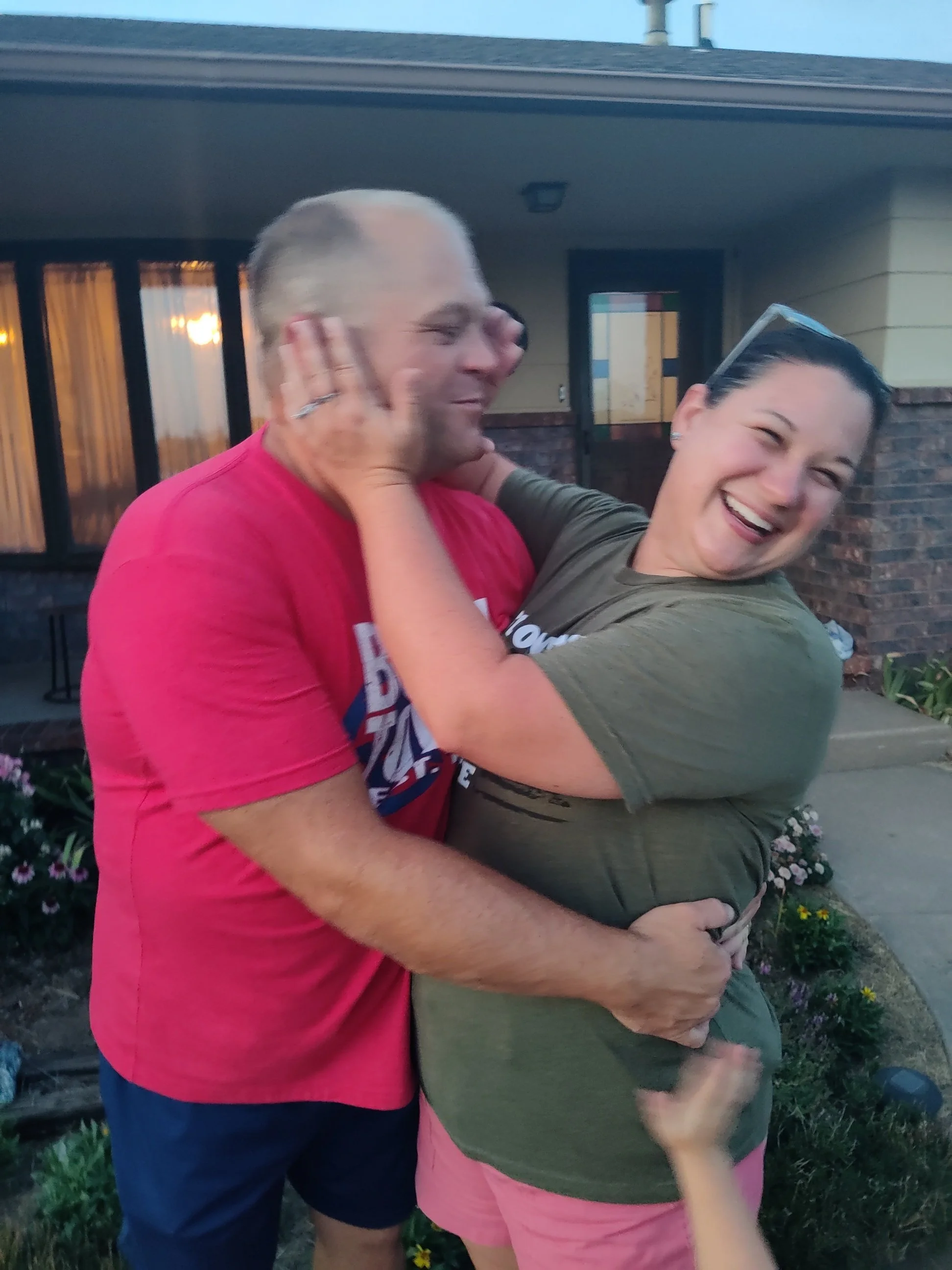A man and a woman are hugging and smiling outdoors, with the man wearing a pink shirt and the woman wearing a green shirt and pink shorts.